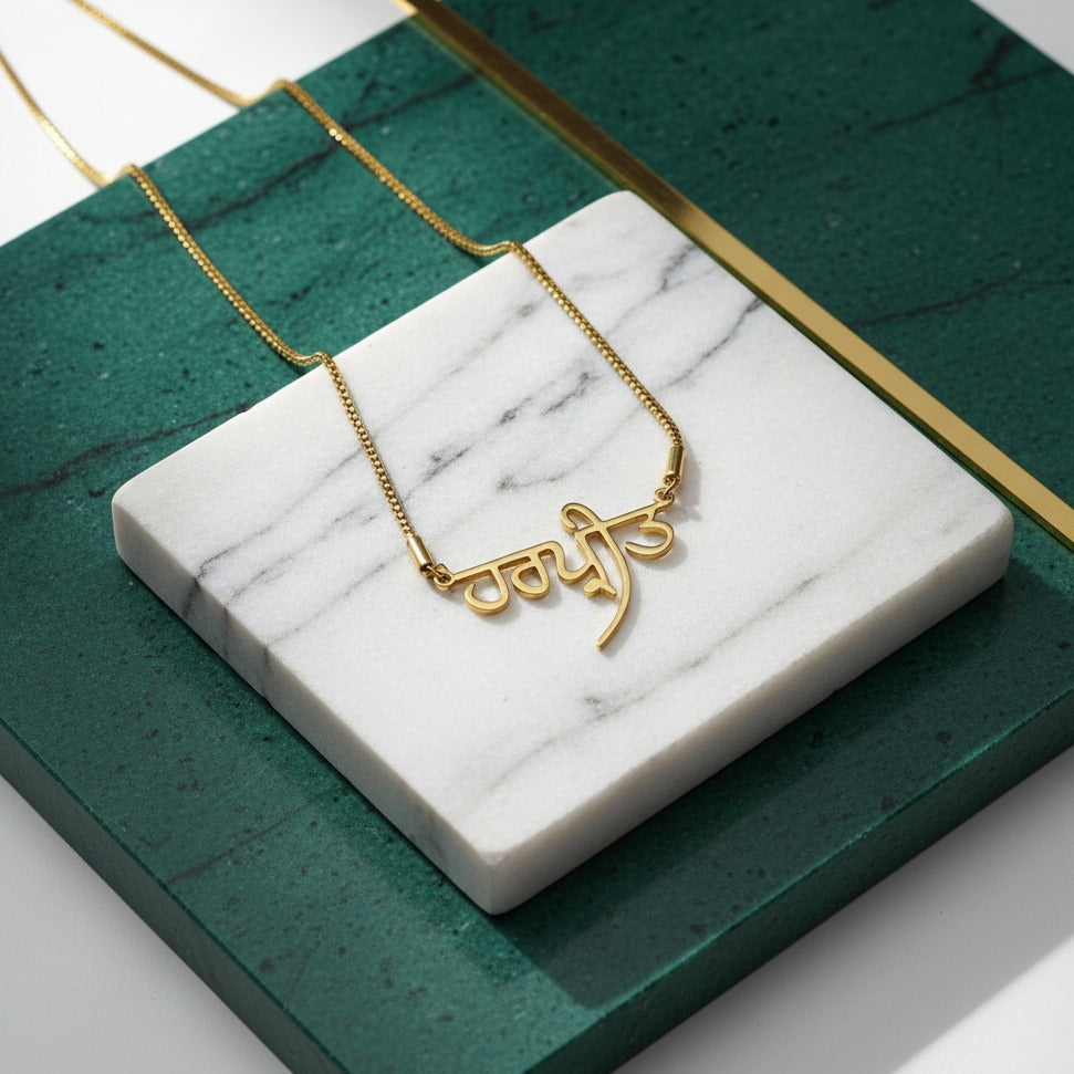 Gold-plated Gurmukhi (Punjabi) name necklace, on a marble tile on green