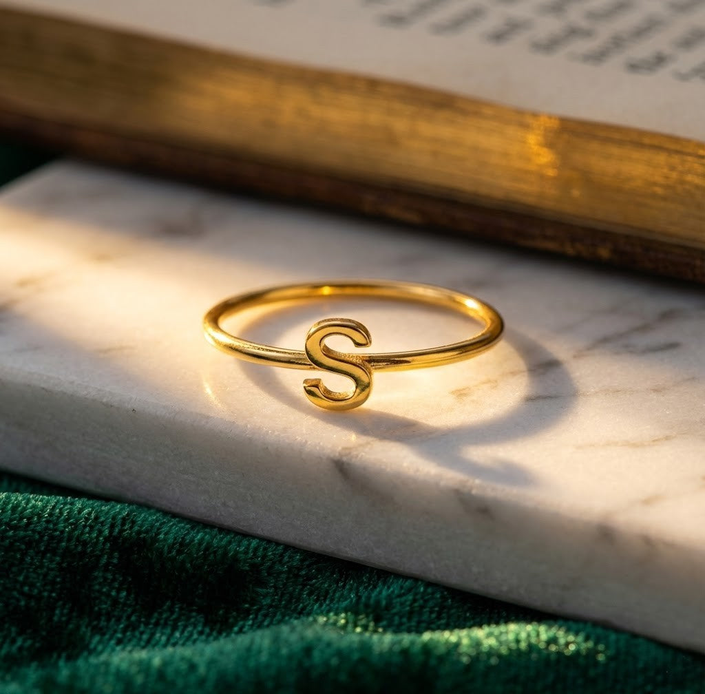 Personalized initial ring, thin gold-tone band with a letter, letter 'S' on marble beside a book