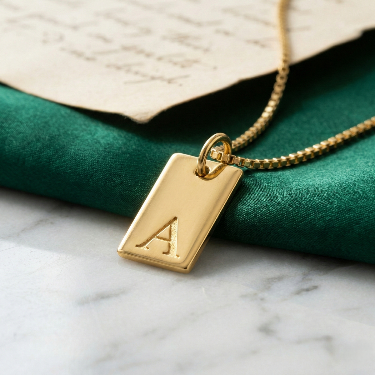 Dainty initial tag necklace, gold-tone rectangular tag with letter 'A', close-up on green velvet with a handwritten note