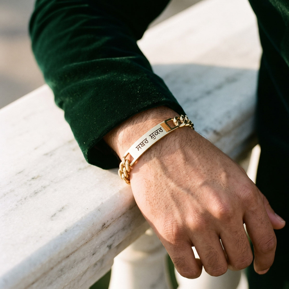 Gold curb name bracelet with ਸਬਰ ਸ਼ੁਕਰ on a man's wrist, resting on a marble railing