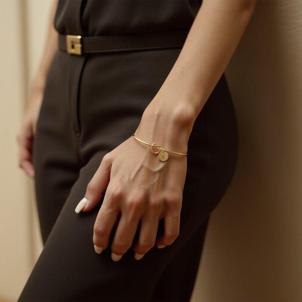 Knotted initial bracelet: open gold-tone bangle with a love knot and round initial charm, worn on a wrist with a black outfit