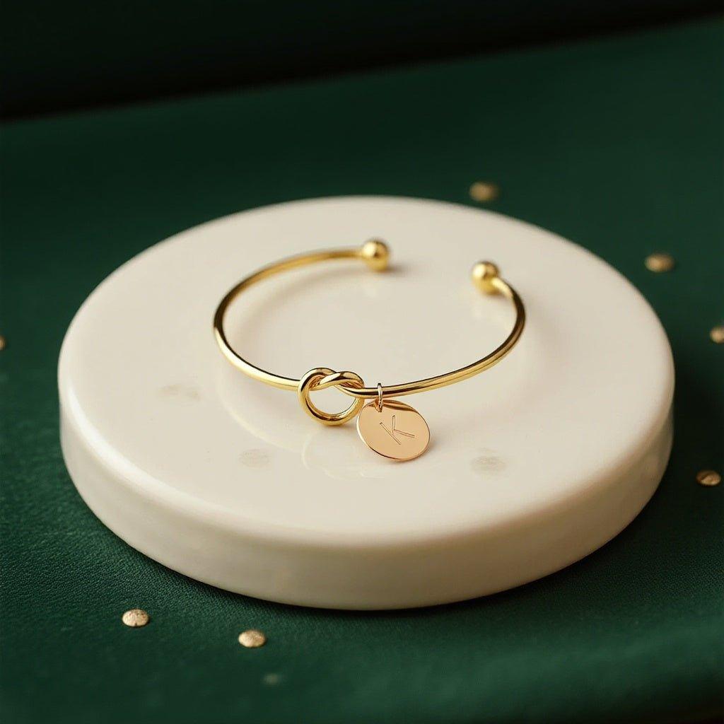 Knotted initial bracelet: open gold-tone bangle with a love knot and round initial charm, on a marble disc with gold flecks