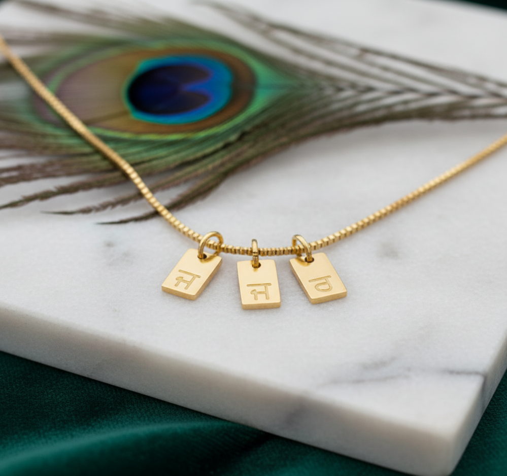 Gold-plated initial tag necklace with Gurmukhi letters, three tags on marble with a peacock feather