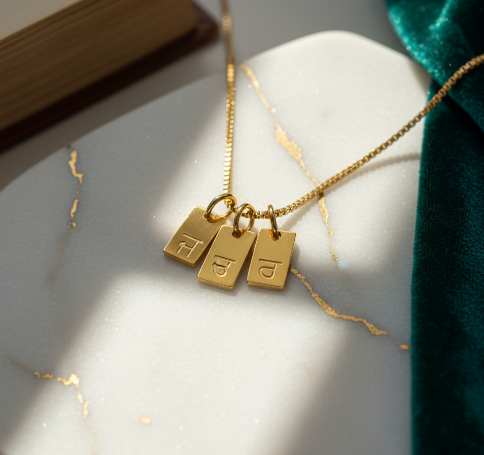 Gold-plated initial tag necklace with Gurmukhi letters, three tags on marble with gold veins