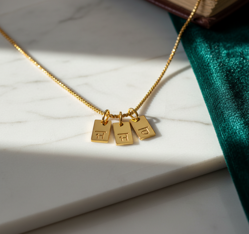 Gold-plated initial tag necklace with Gurmukhi letters, three tags on marble with a book