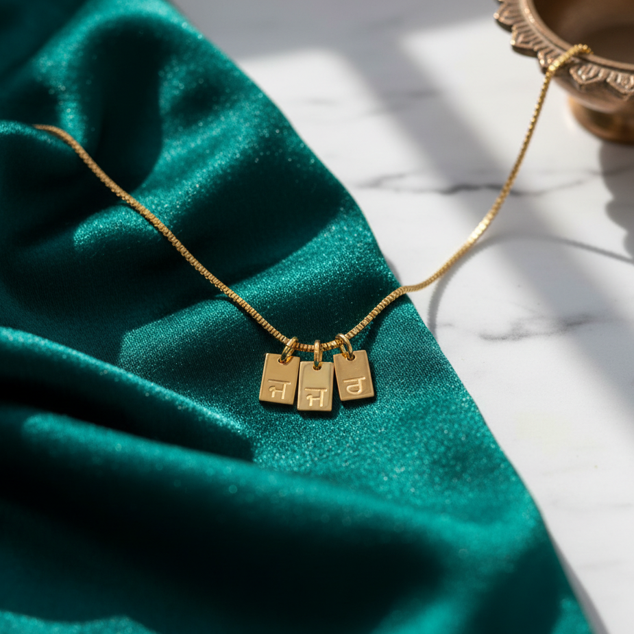 Gold-plated initial tag necklace with Gurmukhi letters, three tags on green velvet