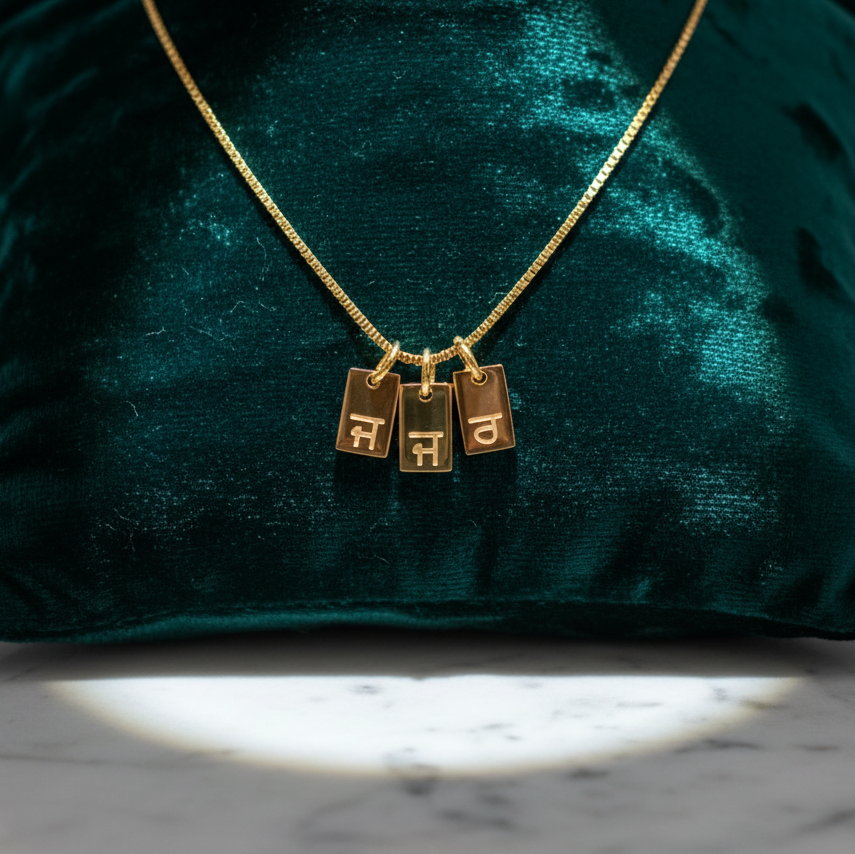 Gold-plated initial tag necklace with Gurmukhi letters, three tags on velvet, darker light