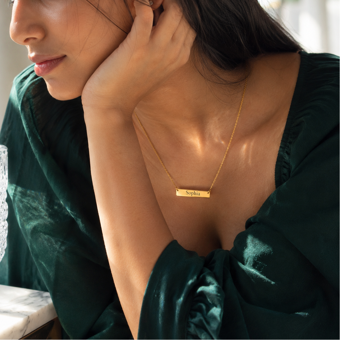 Woman wearing a gold necklace with a nameplate, sitting indoors.