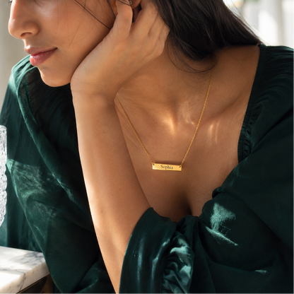 Woman wearing a gold necklace with a nameplate, sitting indoors.