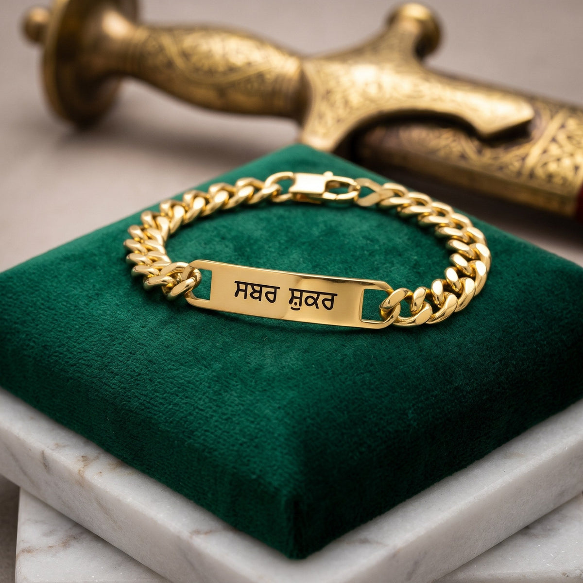 Gold curb name bracelet engraved with ਸਬਰ ਸ਼ੁਕਰ in Punjabi, closed on a green velvet cushion