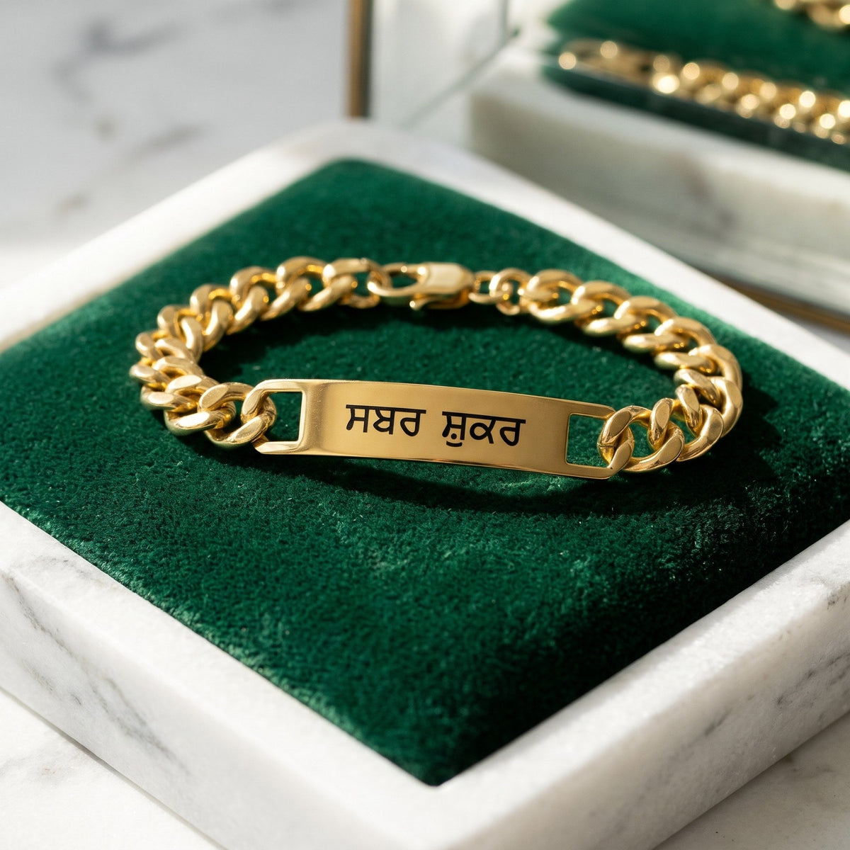 Gold curb name bracelet with ਸਬਰ ਸ਼ੁਕਰ closed in a marble tray with green velvet