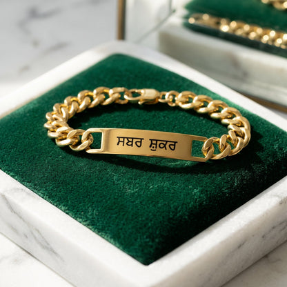 Gold curb name bracelet with ਸਬਰ ਸ਼ੁਕਰ closed in a marble tray with green velvet