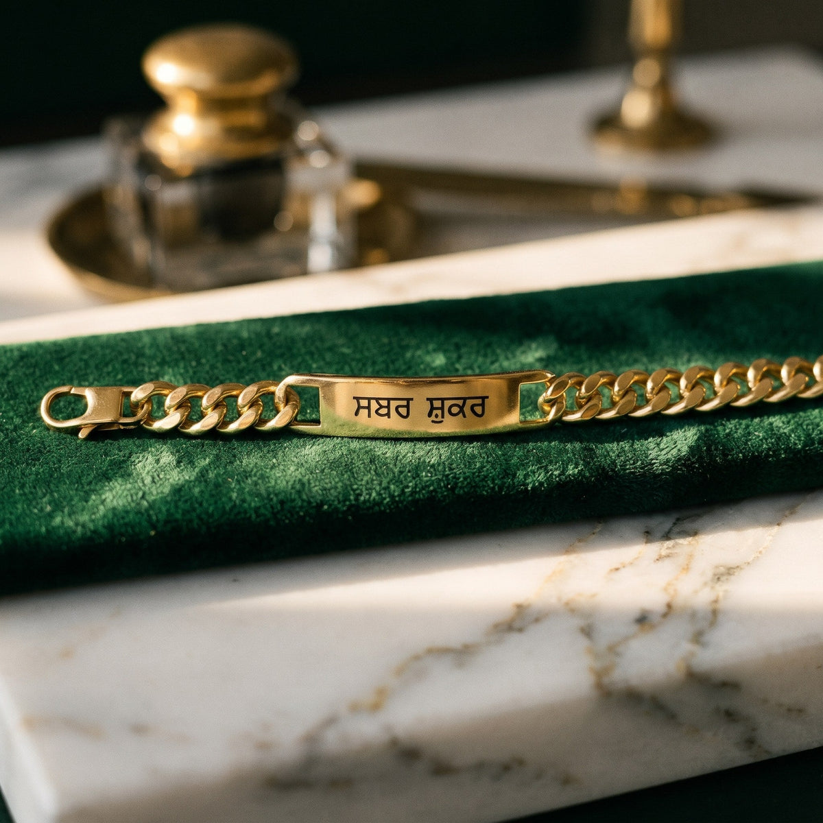Gold curb name bracelet with ਸਬਰ ਸ਼ੁਕਰ on green velvet next to a brass inkwell