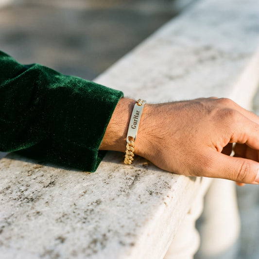 Gold curb name bracelet with GurHar on a man's wrist, resting on a marble railing