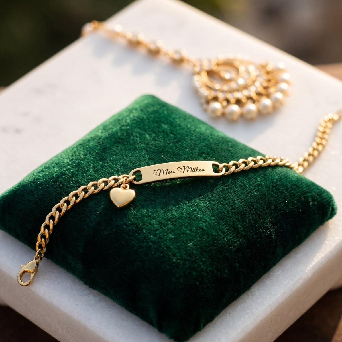 Gold name bracelet with a heart charm, engraved Meri Mithoo, on a green velvet cushion