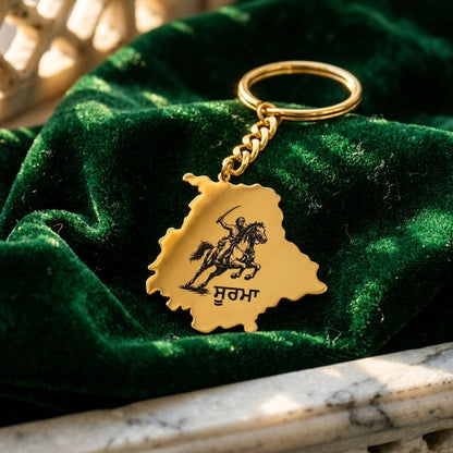 Punjab map gold keychain with the Soorma design and the word ਸੂਰਮਾ, on green velvet